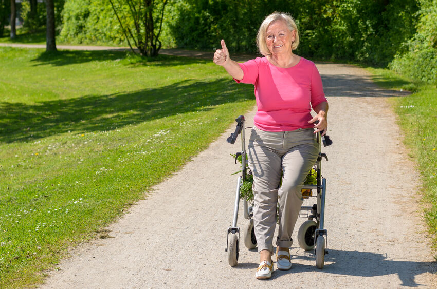 Elderly woman wearing pink blouse sits in wheeled walker on park path while smiling and holding a thumbs up