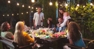 Happy multi-ethnic friends chat at garden in evening. Caucasian man gives high five to girl. African American girl sitting at table. Asian woman laughing. Friends raising and clinking glasses